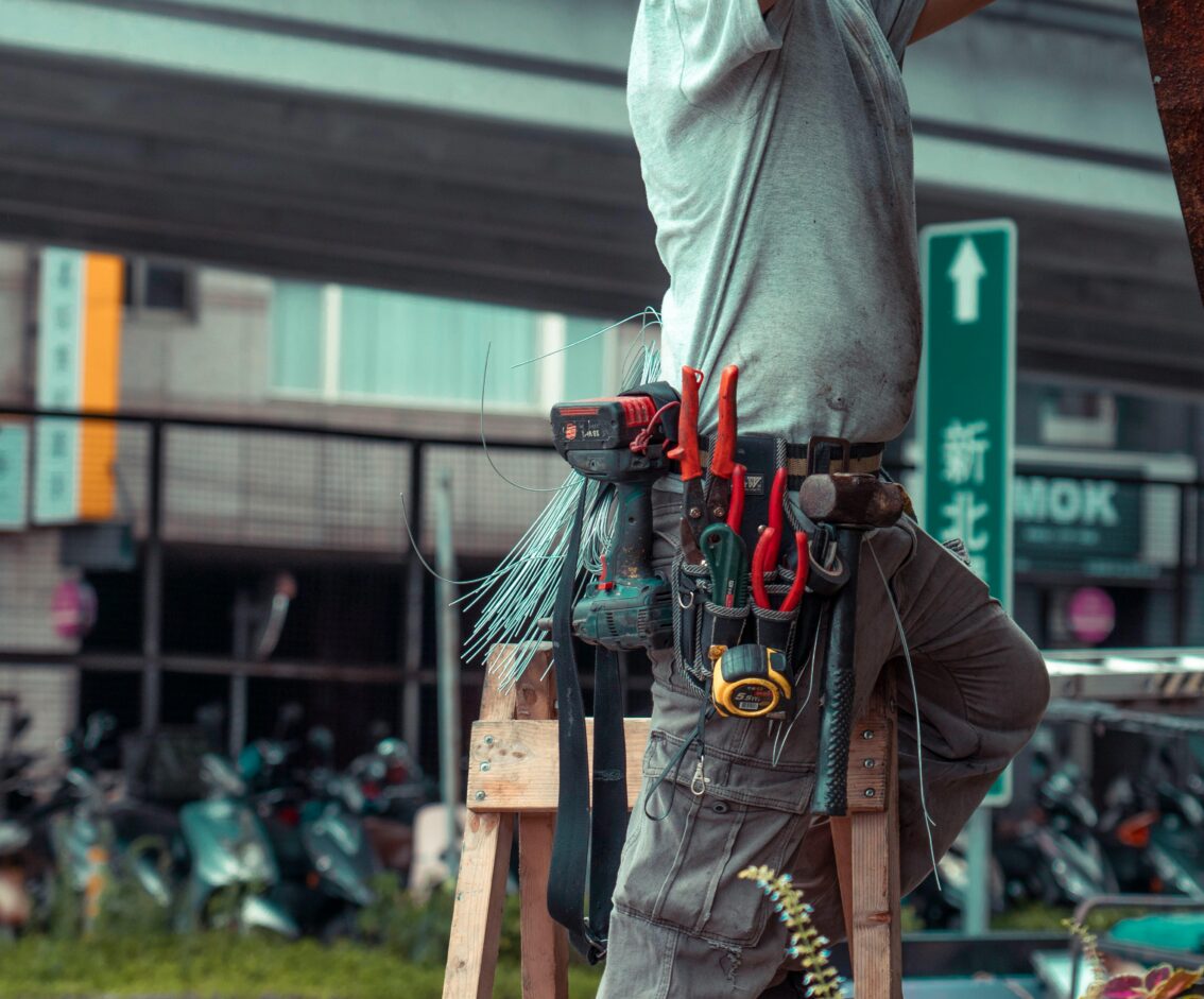 Handyman standing on a ladder with tool belt, repairing outdoors with visible tools.